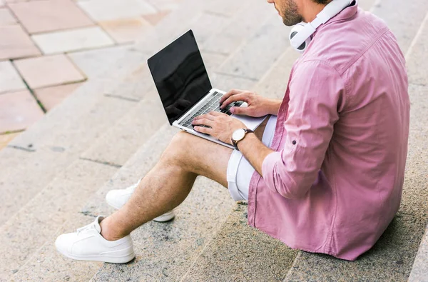 Colpo ritagliato di uomo con le cuffie sul collo utilizzando il computer portatile mentre seduto su gradini sulla strada — Foto stock