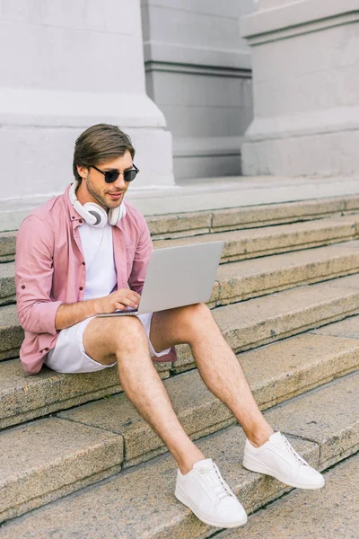 Homem em óculos de sol com fones de ouvido no pescoço usando laptop enquanto sentado em passos na rua — Fotografia de Stock