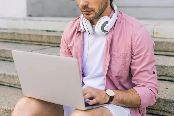 Tiro recortado de homem com fones de ouvido no pescoço usando laptop enquanto sentado em passos na rua — Fotografia de Stock