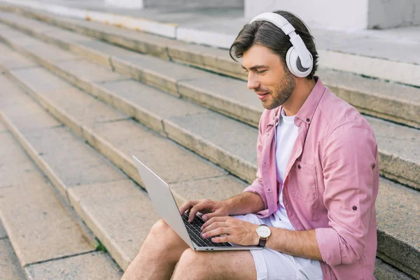 Side view of stylish man in headphones using laptop while sitting on steps on street — Stock Photo