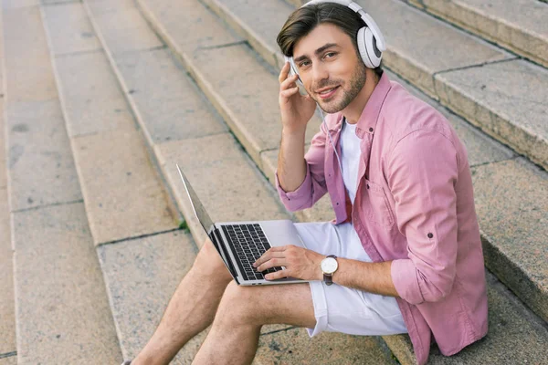 Vista lateral del hombre con estilo en los auriculares con el ordenador portátil sentado en los pasos en la calle - foto de stock