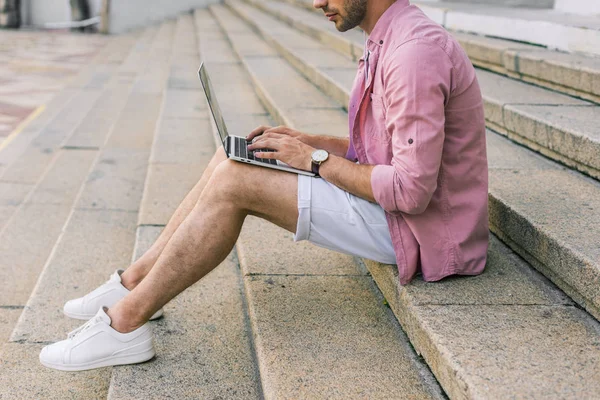 Vista parcial del hombre usando el ordenador portátil mientras está sentado en los escalones de la calle - foto de stock