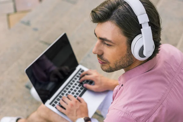 Selective focus of man in headphones with laptop sitting on steps on street — Stock Photo