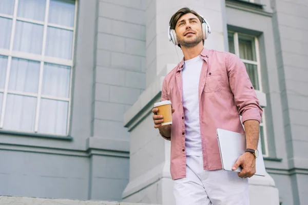 Low angle view of young man in headphones with laptop and coffee to go on street — Stock Photo
