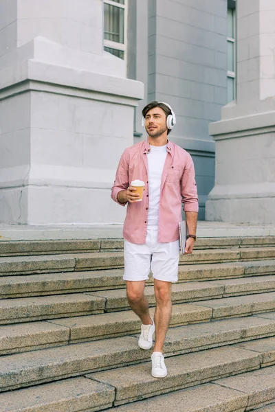 Jeune homme dans les écouteurs avec ordinateur portable et café pour aller dans la rue — Photo de stock