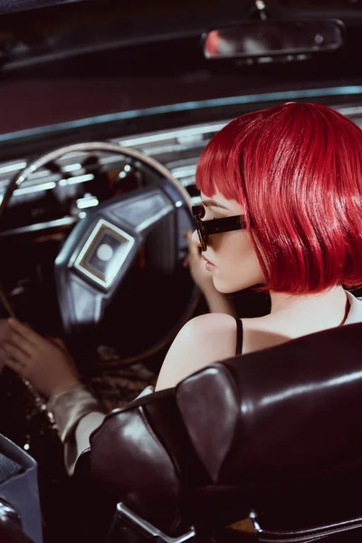 Молодая женщина в красном парике и солнцезащитных очках за рулем ретро автомобиля — стоковое фото