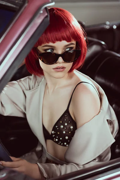 Belle jeune femme en lunettes de soleil, soutien-gorge et trench coat regardant loin tout en étant assis dans la voiture — Photo de stock