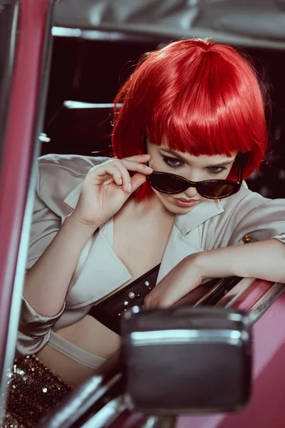 Belle jeune femme en perruque rouge ajuster les lunettes de soleil et regarder miroir de voiture — Photo de stock