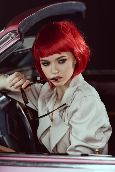 Belle jeune femme en perruque rouge tenant des lunettes de soleil et regardant loin tout en étant assis dans la voiture — Photo de stock