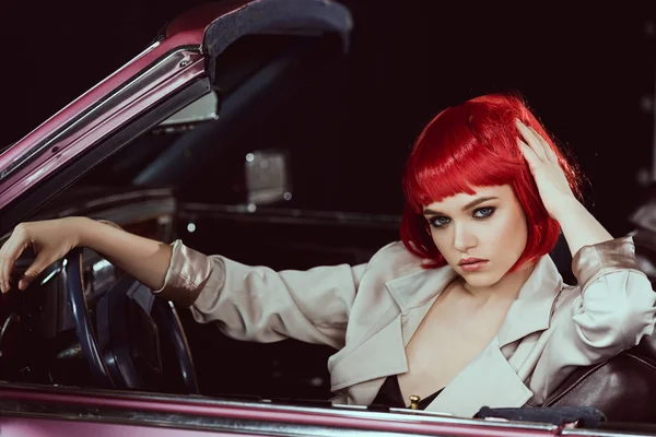 Beautiful young woman in red wig and stylish trench coat sitting in car and looking at camera — Stock Photo