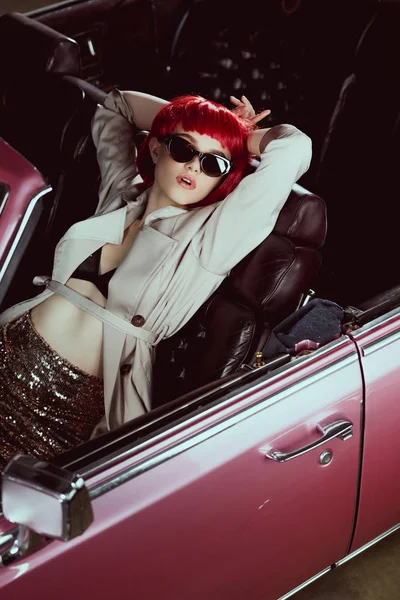 Vue grand angle de la jeune femme élégante en perruque rouge, trench coat et lunettes de soleil assis dans une voiture rétro — Photo de stock