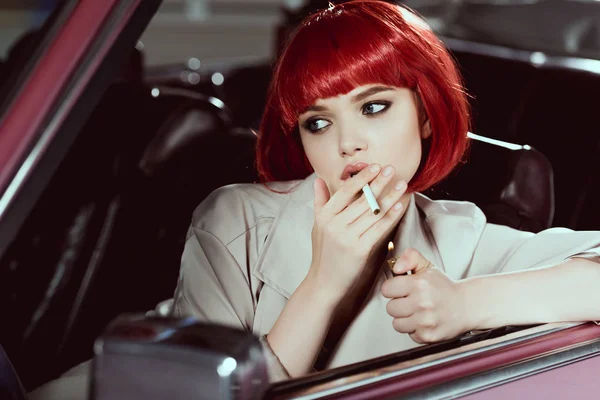 Привлекательная девушка в красном парике курит сигарету и смотрит в сторону, сидя в машине — стоковое фото
