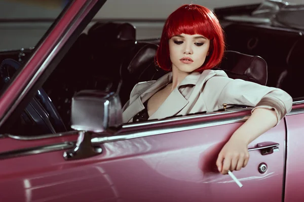 Hermosa chica con estilo en peluca roja sosteniendo el cigarrillo y sentado en coche vintage - foto de stock