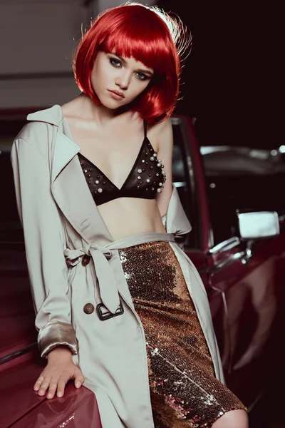 Beautiful young woman in red wig, bra and trench coat sitting on classic car and looking at camera — Stock Photo