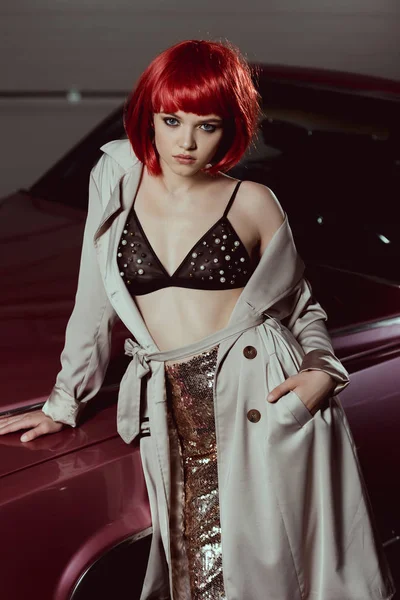 High angle view of stylish girl in trench coat and bra leaning at car and looking at camera — Stock Photo