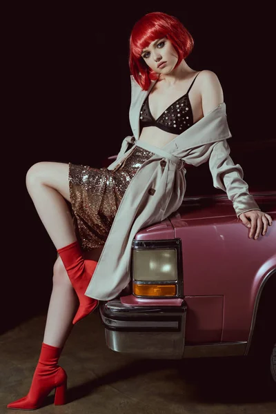 Full length view of beautiful girl in bra and trench coat sitting on car and looking at camera — Stock Photo