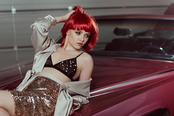 Sensual young woman in bra and trench coat lying on car and looking away — Stock Photo