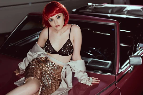 High angle view of beautiful young woman in bra and trench coat sitting on car and looking at camera — Stock Photo