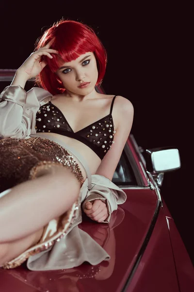 Красивая молодая женщина в бюстгальтере и стильном плаще смотрит в камеру, когда лежит на машине — стоковое фото