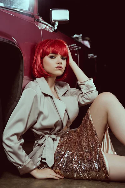 Menina elegante em peruca vermelha olhando para a câmera enquanto sentado perto do carro vintage — Fotografia de Stock