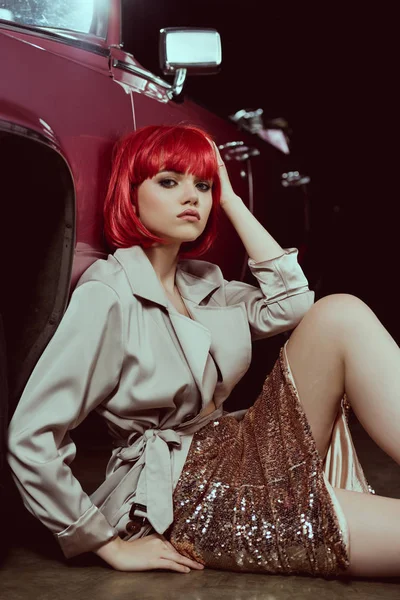 Stylish girl in red wig looking at camera while sitting near vintage car — Stock Photo