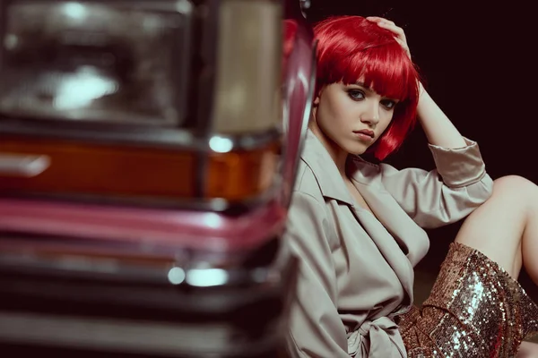 Selektiver Fokus der schönen Frau mit roter Perücke, die in die Kamera schaut, während sie in der Nähe des Retro-Autos sitzt — Stockfoto
