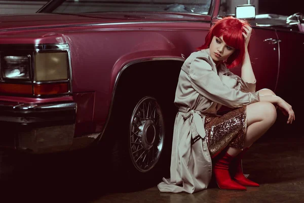 Hermosa mujer joven con estilo mirando hacia otro lado mientras se agacha cerca de coche vintage - foto de stock