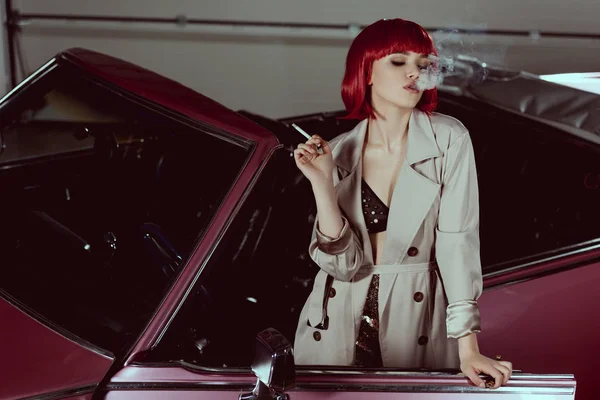 Стильна дівчина в червоній перуці і тренч пальто куріння сигарети в класичному автомобілі — стокове фото