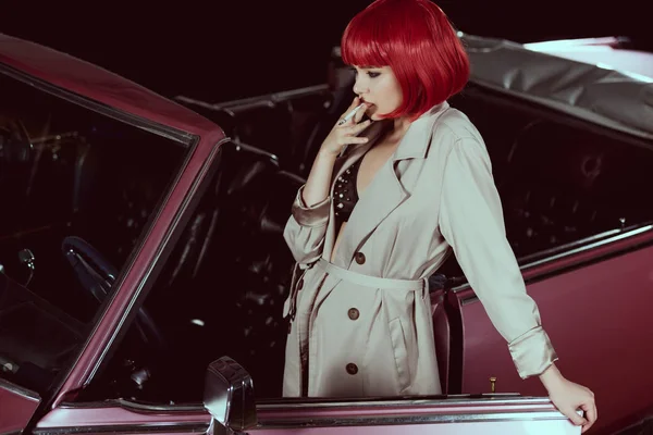 Vista de ángulo alto de la chica con estilo en peluca roja y gabardina fumar cigarrillo cerca de coche vintage - foto de stock