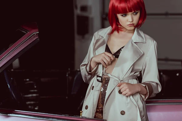 Stylish girl in red wig tying trench coat and looking away near car — Stock Photo