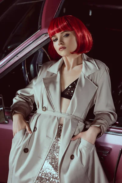 Красивая стильная девушка в бюстгальтере и пальто, смотрящая в камеру, опираясь на автомобиль — стоковое фото