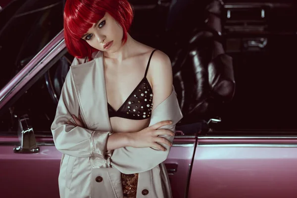 Hermosa chica elegante en sujetador y gabardina mirando a la cámara mientras está de pie con los brazos cruzados en el coche - foto de stock