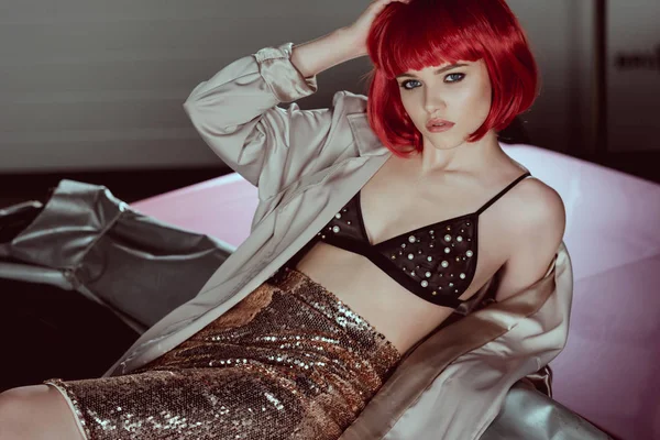 Beautiful young woman in red wig, bra and trench coat looking at camera while sitting in car — Stock Photo