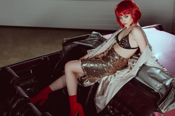Vista de ángulo alto de la chica con estilo en peluca roja y gabardina mirando a la cámara mientras está sentado en el coche retro — Stock Photo