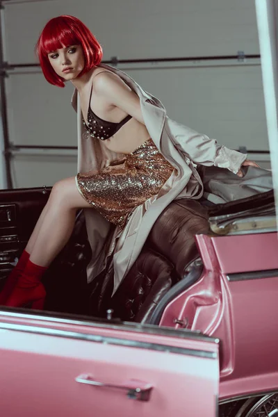 Young woman in trench coat, glittering skirt and bra looking away while sitting on retro car — Stock Photo