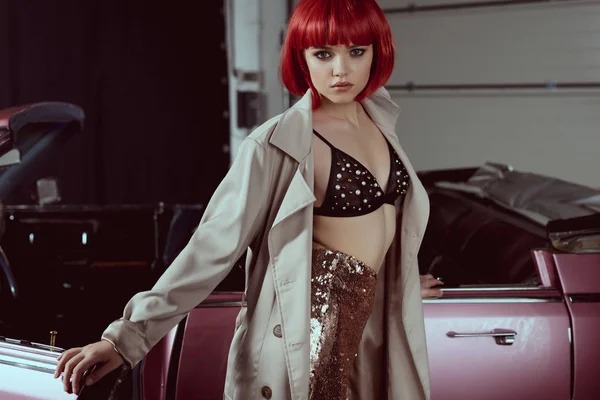 Красивая молодая женщина в модном плаще смотрит в камеру, стоя рядом с ретро-машиной — стоковое фото