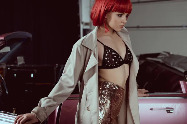 Beautiful young woman in fashionable trench coat looking away while standing near retro car — Stock Photo