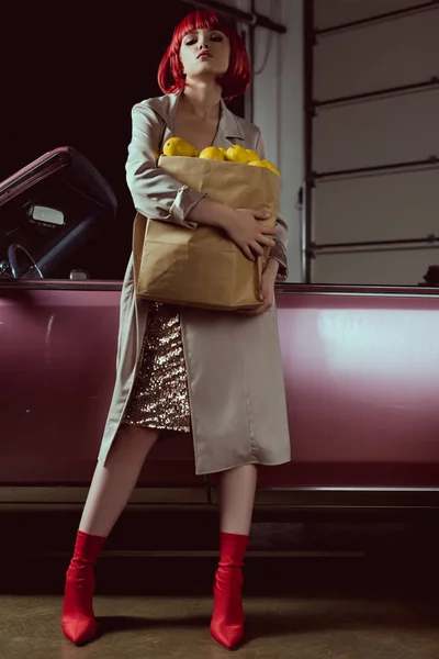 Vue à angle bas de belle femme blonde en perruque rouge et élégant trench coat tenant sac en papier avec des citrons et en regardant la caméra tout en se tenant près de la voiture classique — Photo de stock