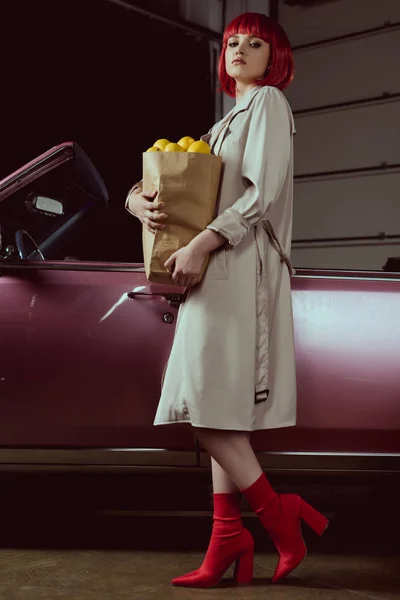 Vue à angle bas de jeune femme à la mode tenant sac en papier avec des citrons et regardant la caméra tout en se tenant près de la voiture rétro — Photo de stock