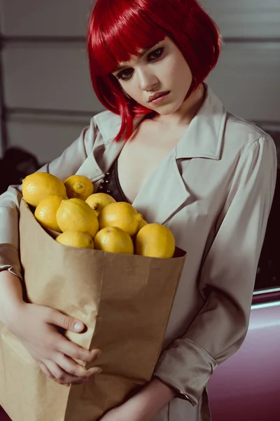 Pensive girl in red wig and stylish trench coat holding paper bag with lemons — Stock Photo