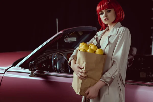 Mujer en peluca roja y elegante gabardina sosteniendo bolsa de papel con limones y mirando a la cámara mientras está de pie cerca de coche retro - foto de stock