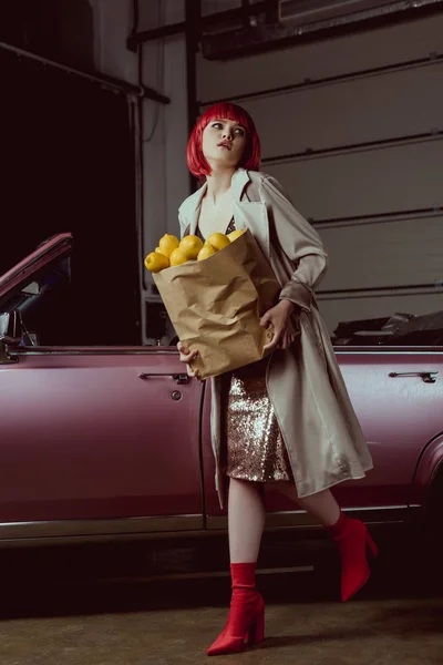 Женщина в красном парике и стильном плаще держа бумажный пакет с лимонами возле ретро автомобиля — стоковое фото
