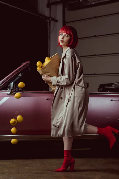 Chica en peluca roja y elegante gabardina sosteniendo bolsa de papel con limones que caen cerca de coche retro - foto de stock