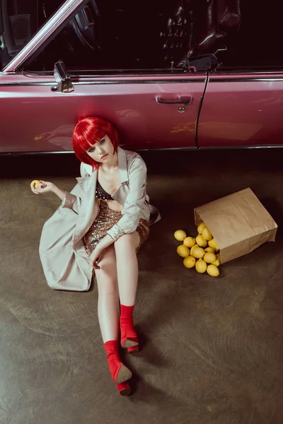 High angle view of beautiful girl in stylish trench coat holding lemon and looking at camera while sitting near retro car — Stock Photo