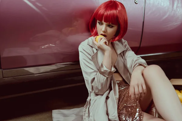 Стильная молодая женщина в красном парике ест лимон, сидя рядом с ретро-машиной — стоковое фото