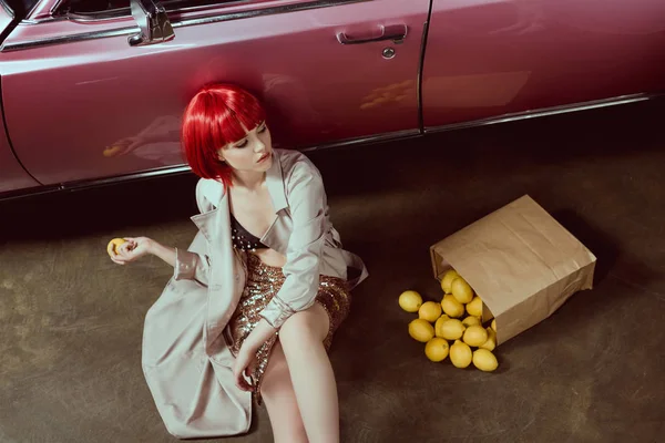 High angle view of beautiful girl in stylish trench coat holding lemon while sitting near retro car — Stock Photo