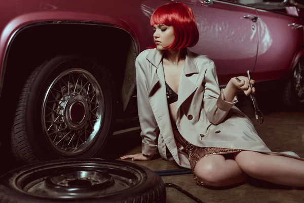 Beautiful young woman in trench coat holding wrench and repairing car — Stock Photo