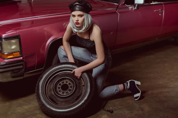 Visão de alto ângulo de bela menina loira elegante em couro boina reparando roda do carro e olhando para a câmera — Fotografia de Stock