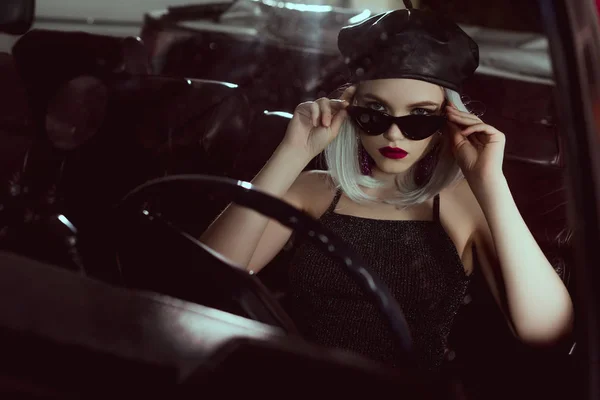 Belle fille blonde à la mode en béret et lunettes de soleil assis dans la voiture et regardant la caméra — Photo de stock