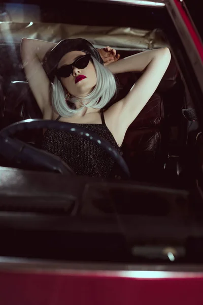 Sensual stylish blonde girl in beret and sunglasses sitting with hands behind head in vintage car — Stock Photo